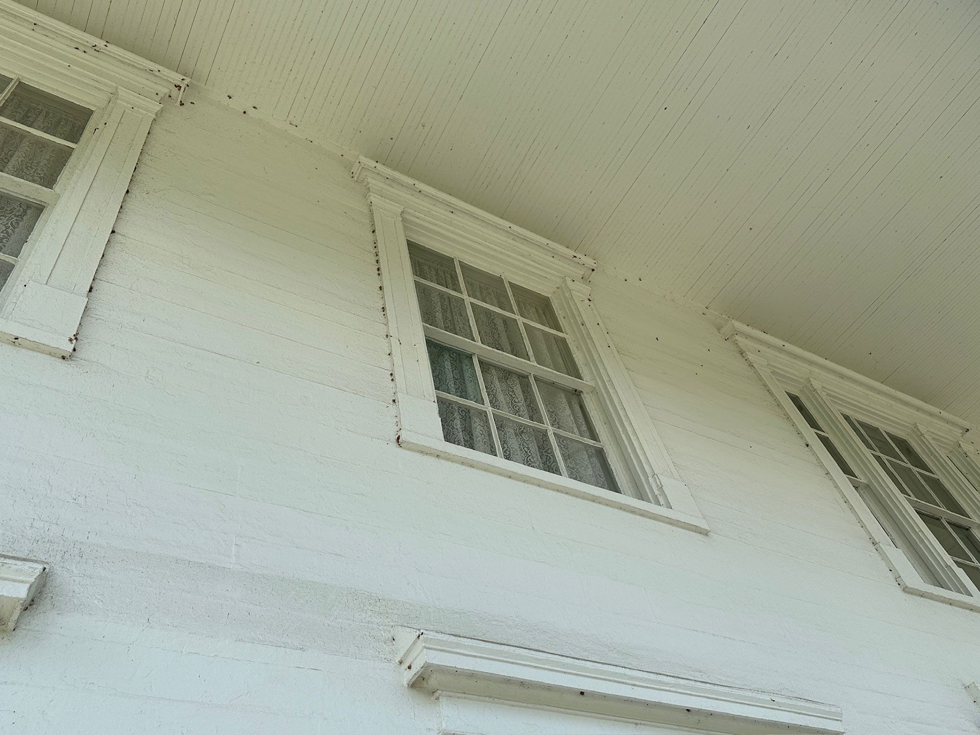 White house exterior with three windows and a shutter, viewed from below against a pale sky