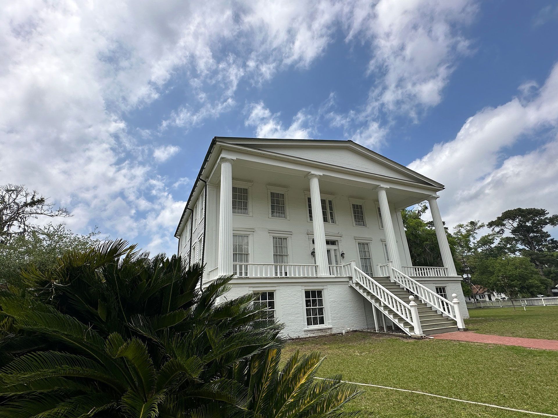 White colonial-style mansion with large columns, front stairs, and a manicured lawn under a cloudy sky