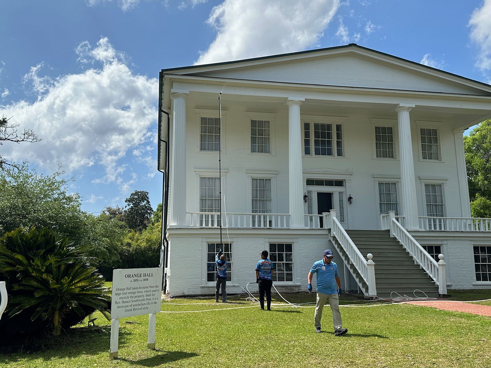 White historic mansion with columns and stairs, three people standing on the lawn under a blue sky.