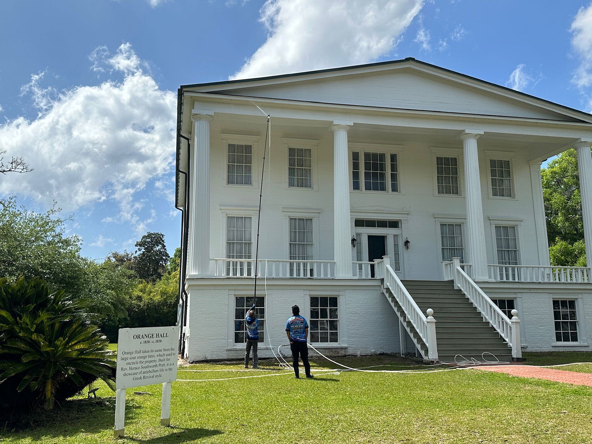 White colonial-style building with tall columns, front stairs, and two people standing on the lawn