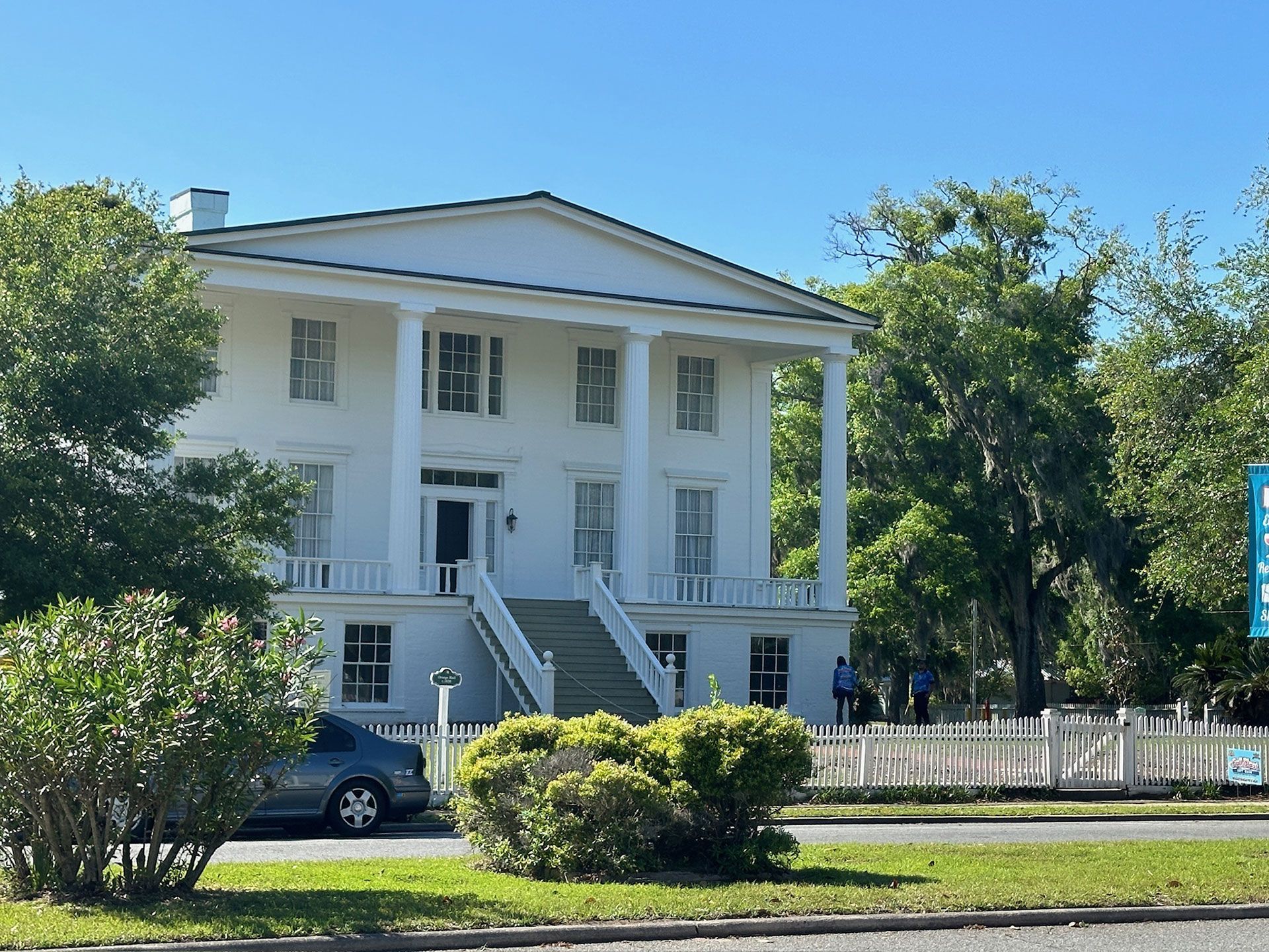 White neoclassical mansion with tall columns, front steps, and trees under a clear blue sky.