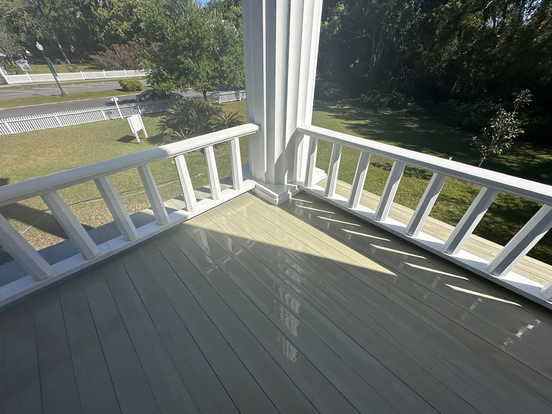 Sunlit porch with white railings and pale gray floorboards overlooking trees and a street