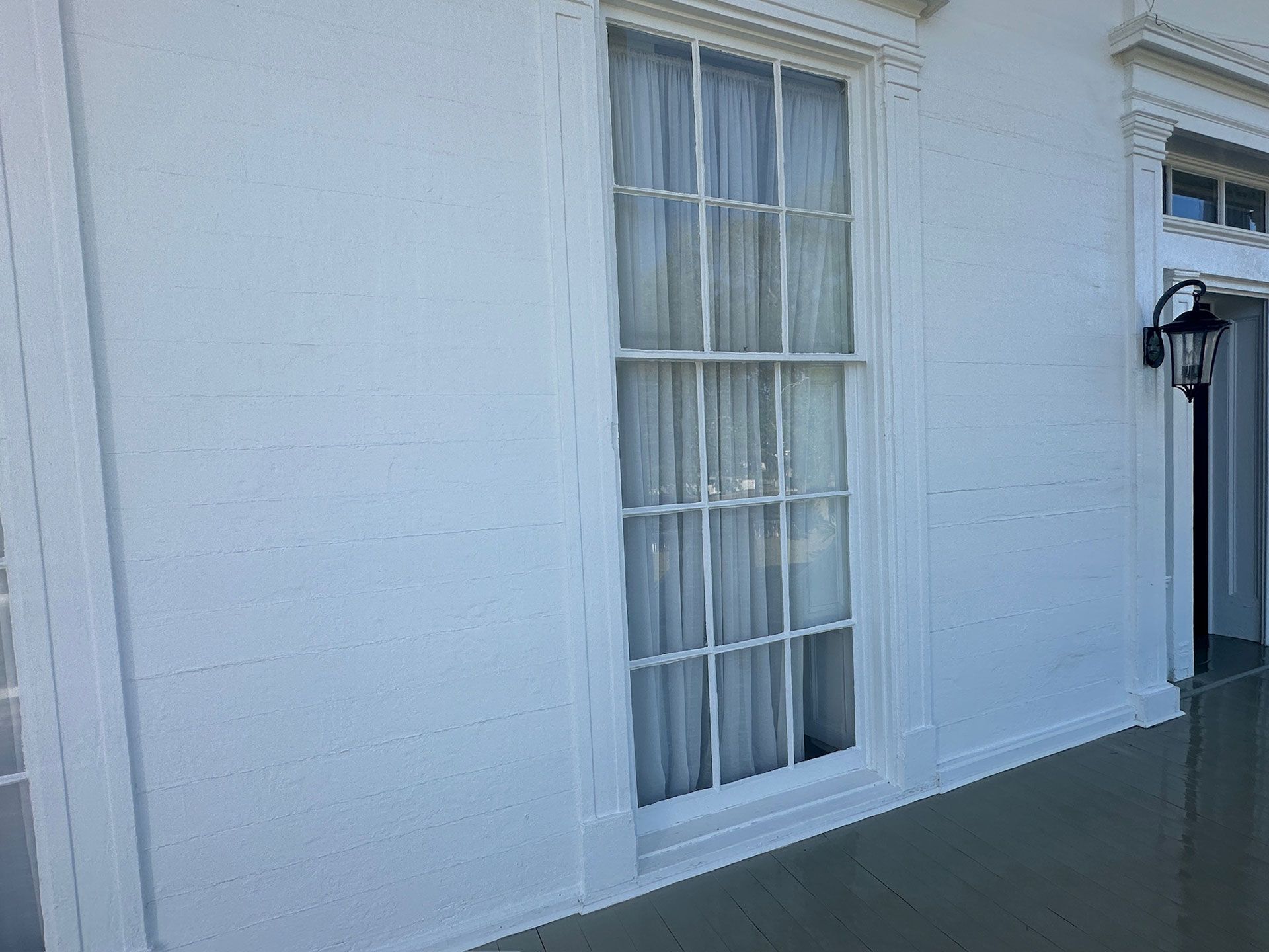 White exterior wall with a tall window and a black wall lantern beside a wet sidewalk