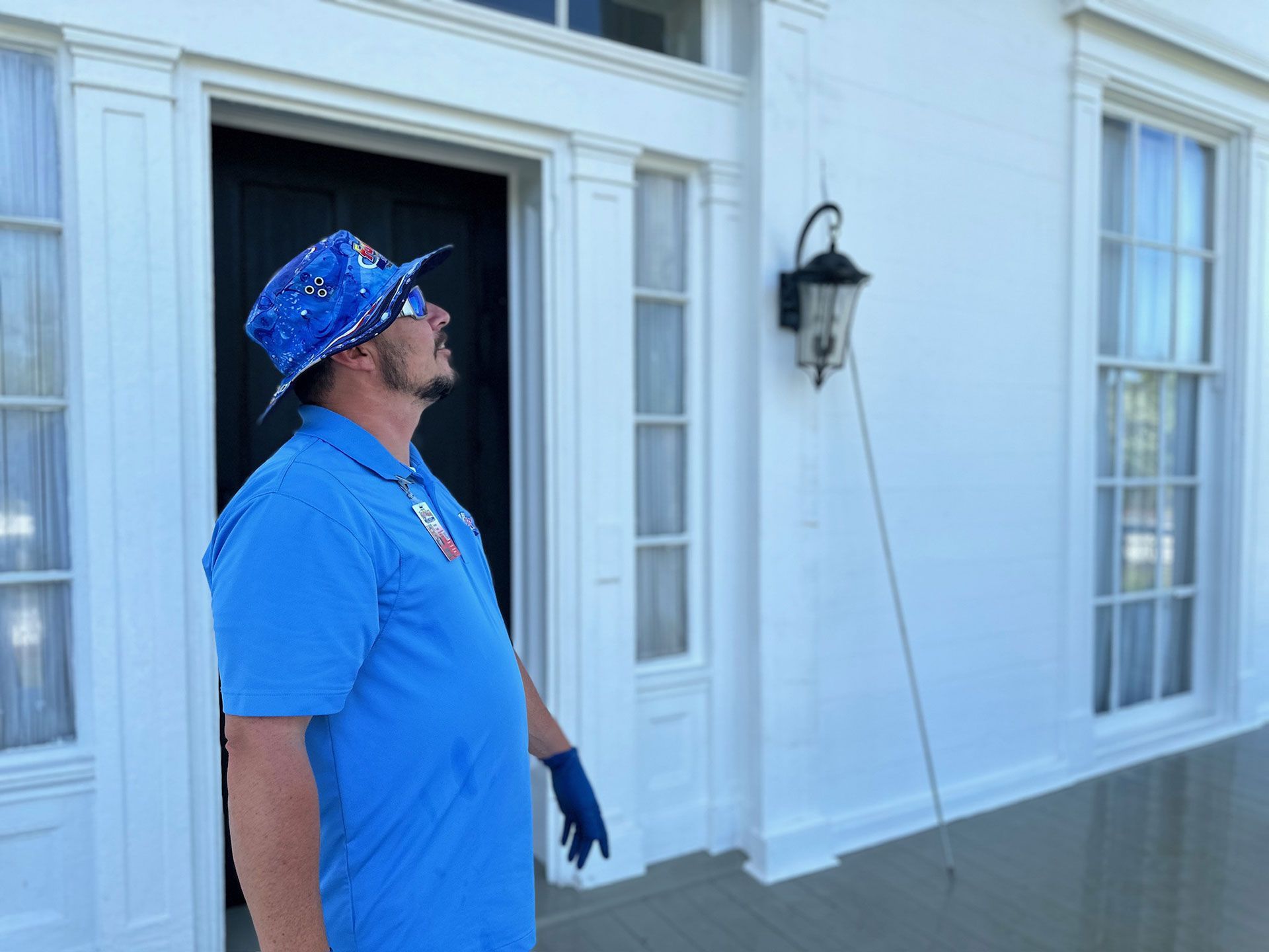 Man in blue shirt and cap stands on a porch outside a white house, looking upward.