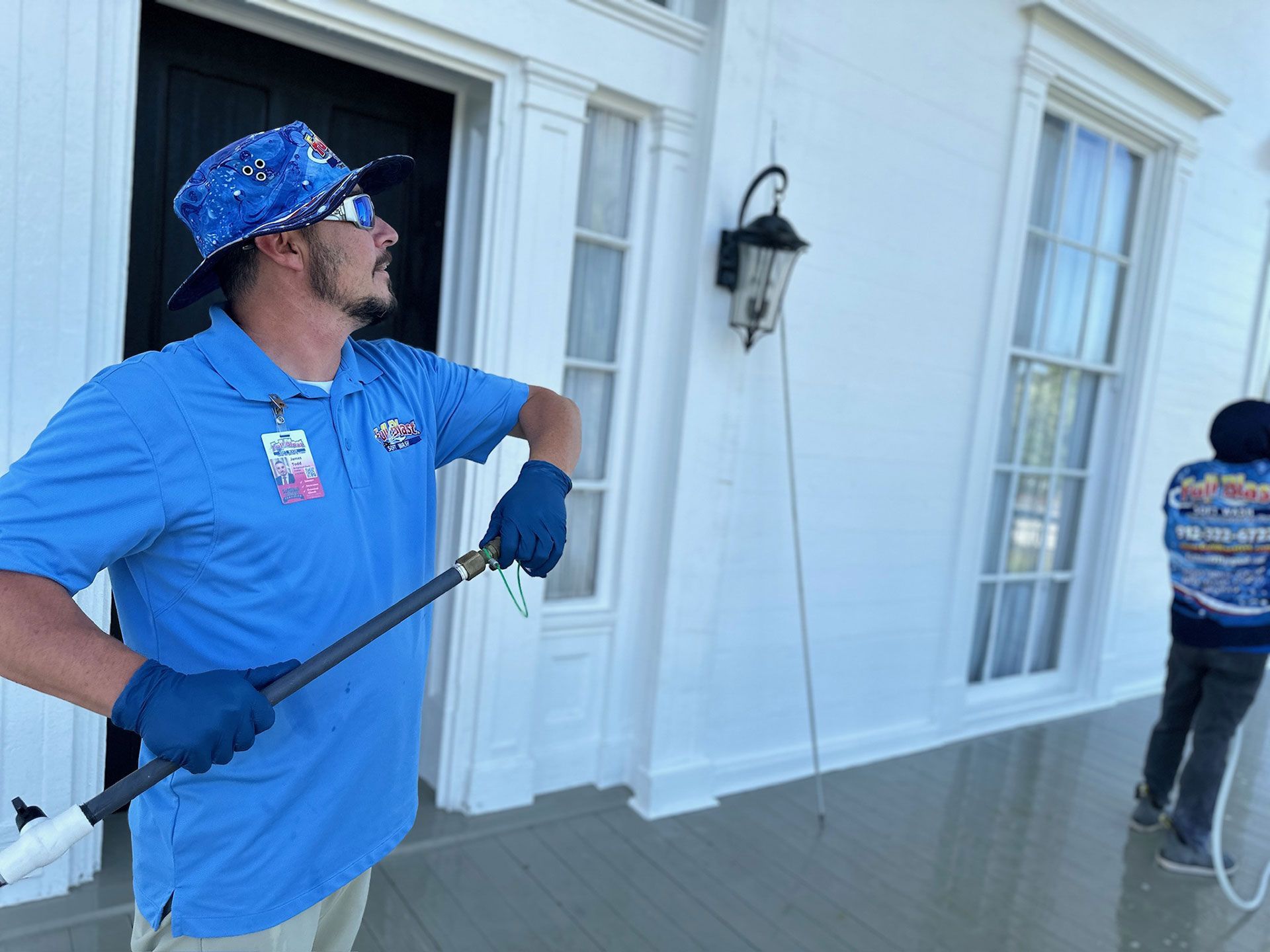 Blue-shirted worker on a porch holds a long-handled tool near white siding, with another worker in the background.