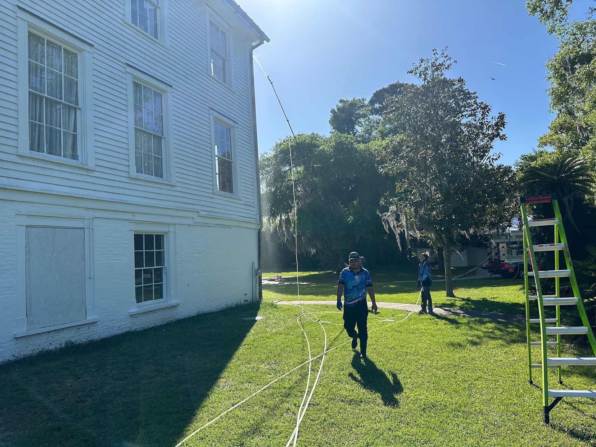 People mowing a sunny lawn beside a white house, with a ladder on the right.