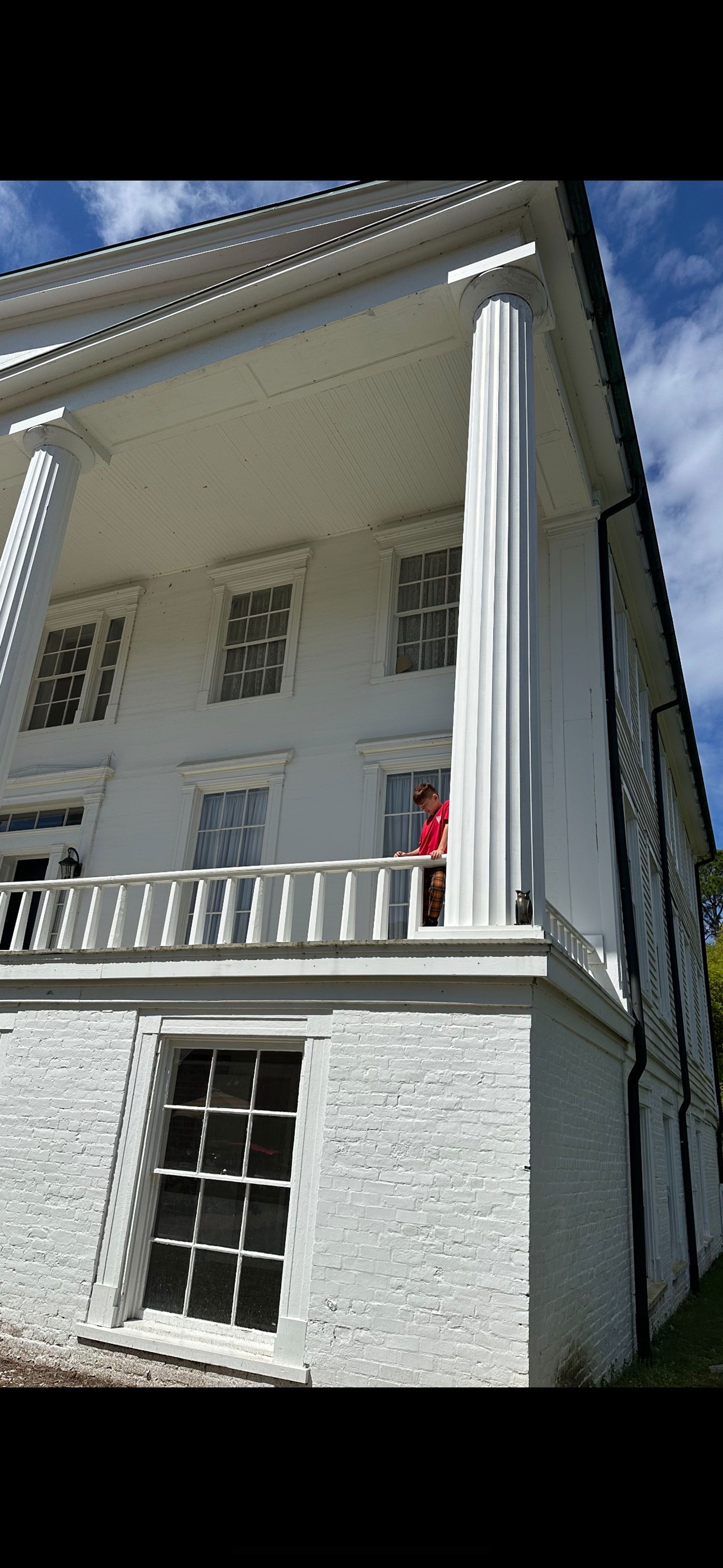 White columned historic house with a balcony and a person in red standing on the porch under a blue sky