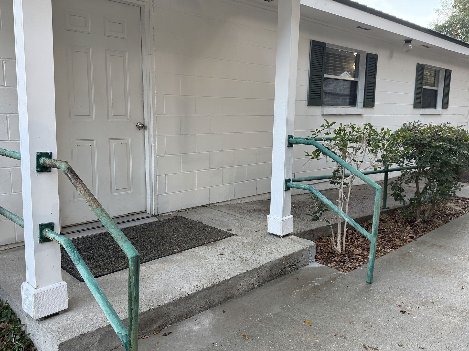 Front entrance with concrete steps, green handrails, and a white door on a small building