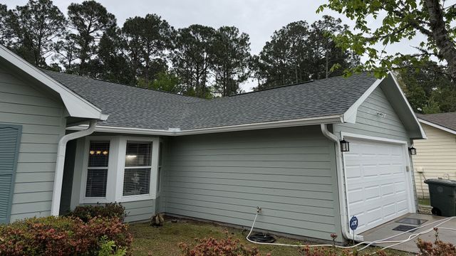 Single-story suburban house with gray siding, white garage door, and a dark shingled roof under cloudy skies