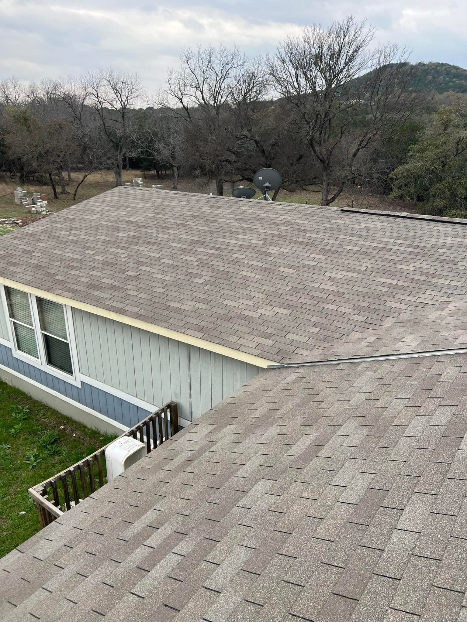 Rooftops of a house with gray shingles, bordered by leafless trees and hills under an overcast sky