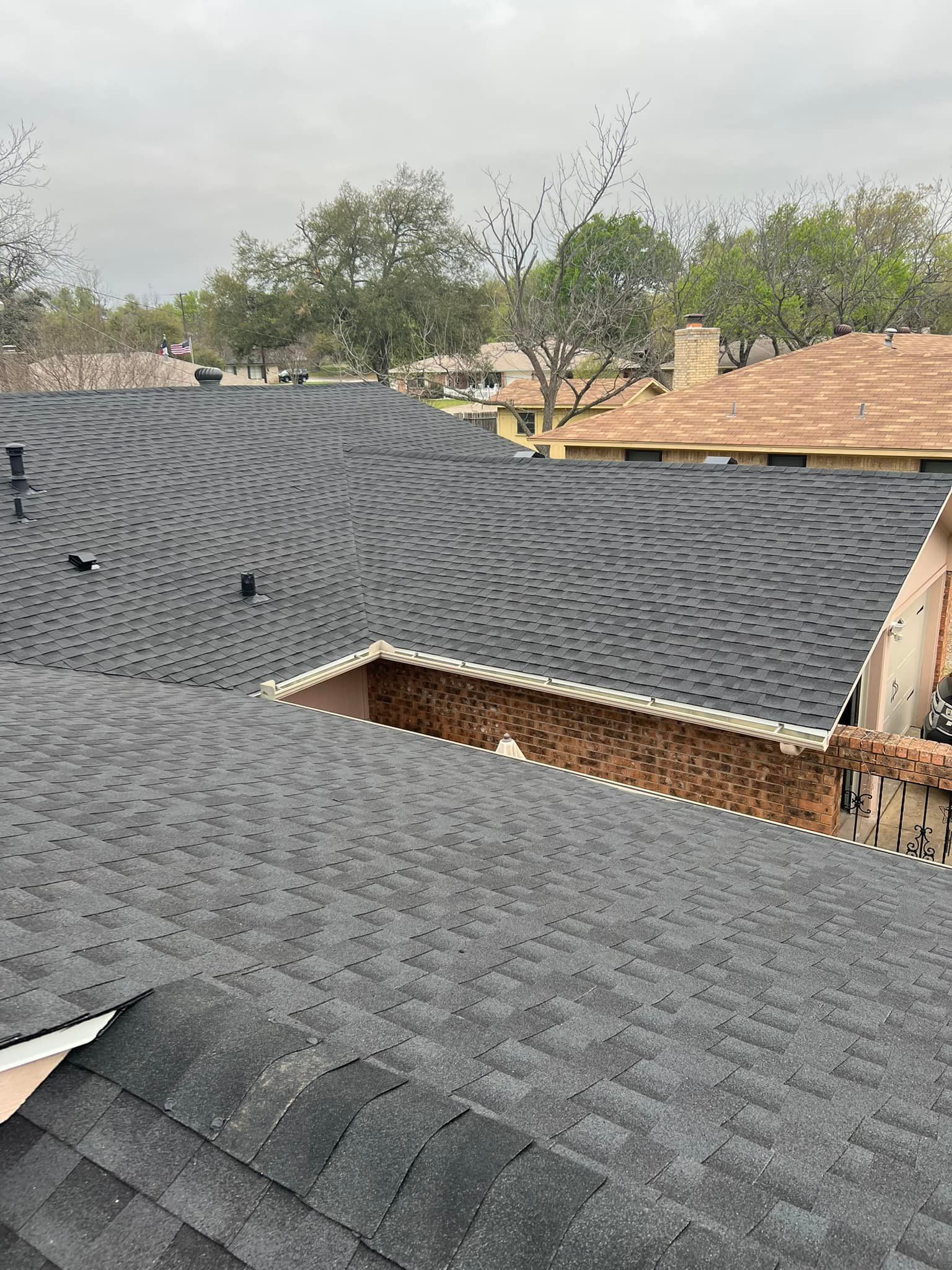 Residential rooftops with dark shingles under a cloudy sky, with trees and neighboring houses in the background