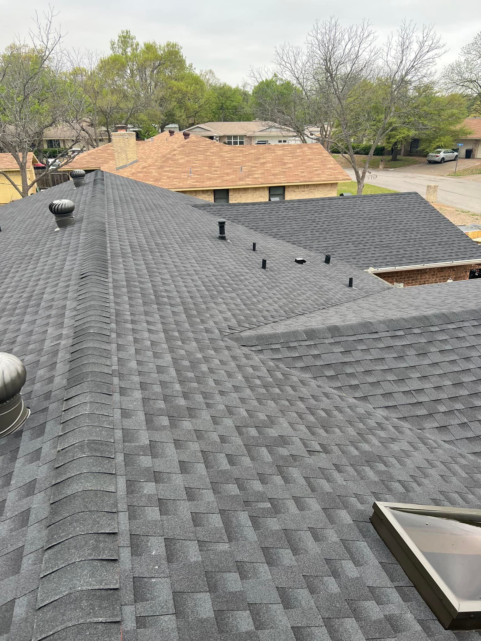 Rooftop view of dark gray shingles with skylights and nearby tan houses under trees