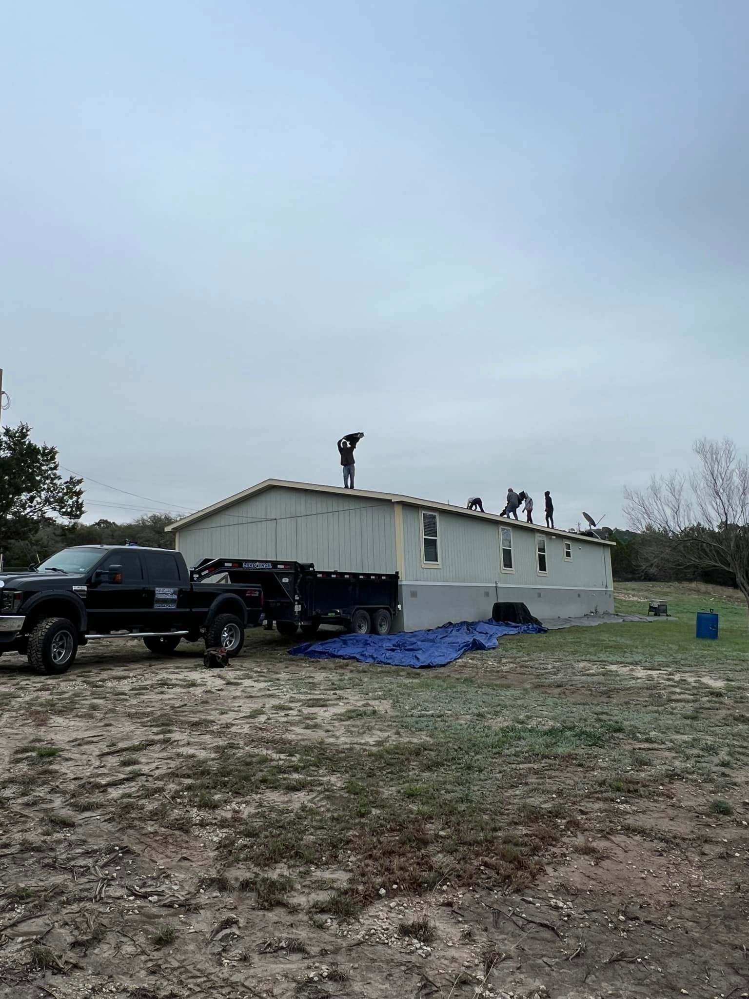 Workers on a white house roof with trucks and a blue tarp in a muddy yard under an overcast sky