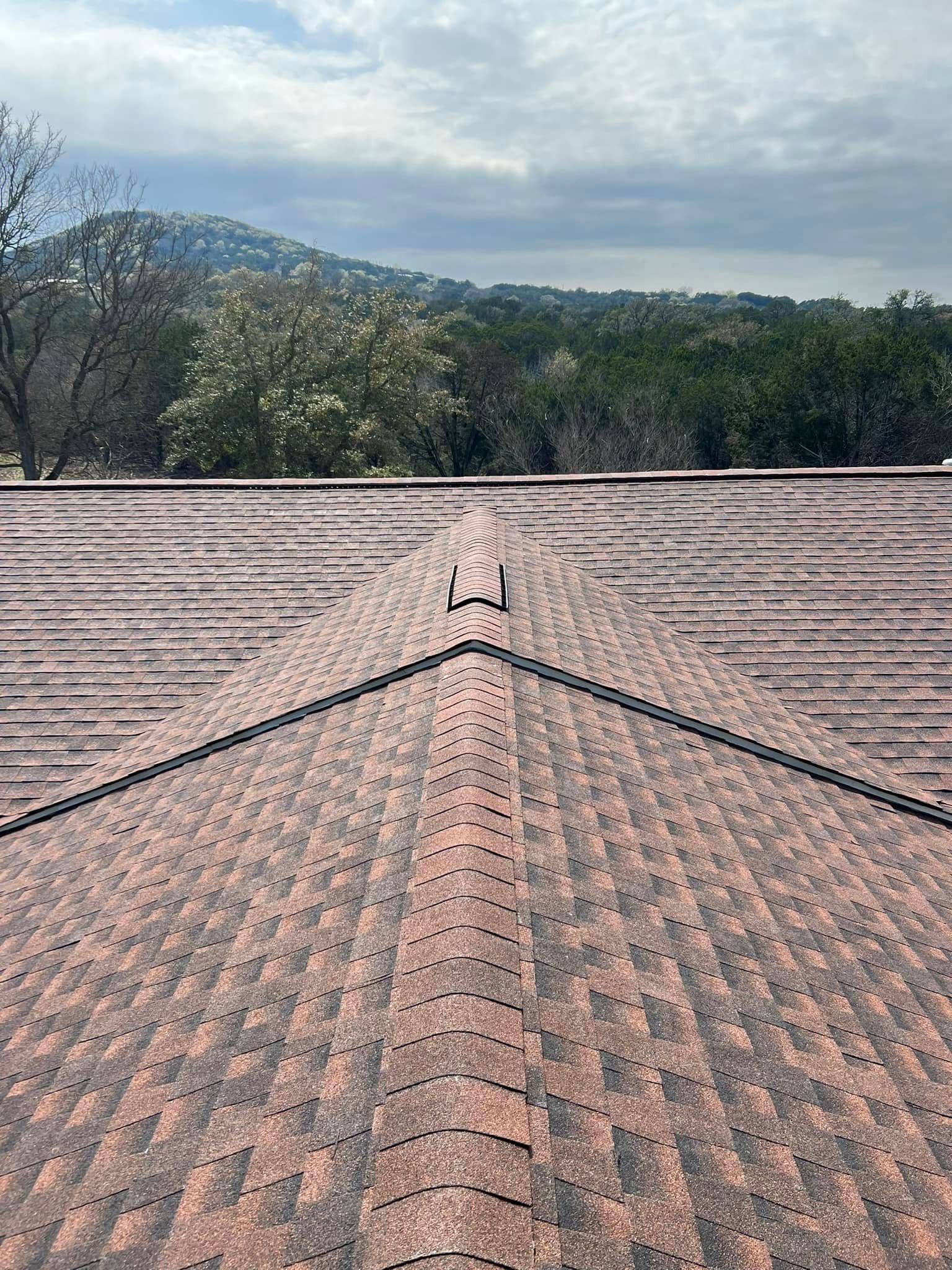 Roof shingles with a central ridge line, overlooking trees and hills under a cloudy sky