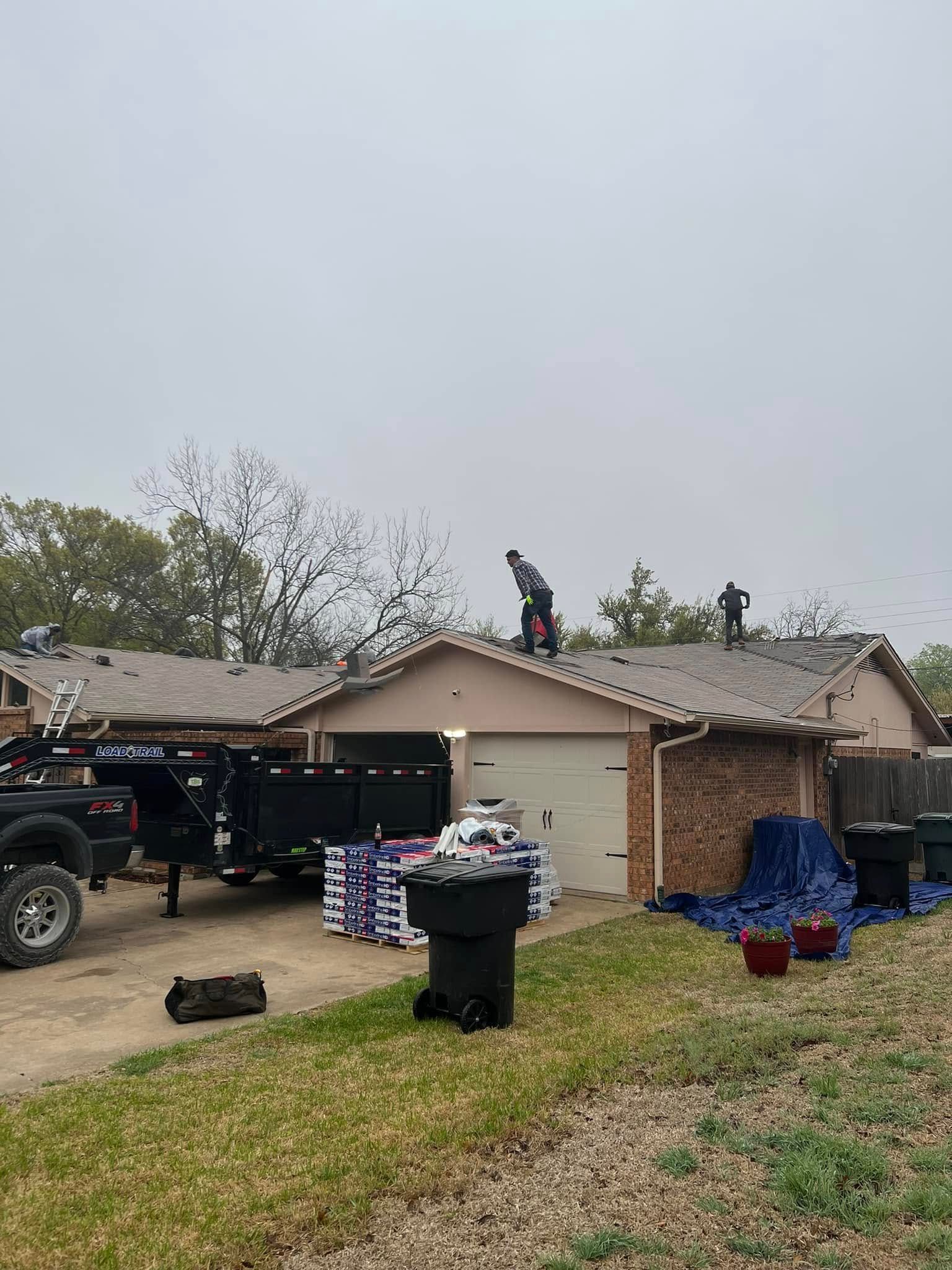 Worker on a roof beside a house with a truck, trash bins, and debris in the yard