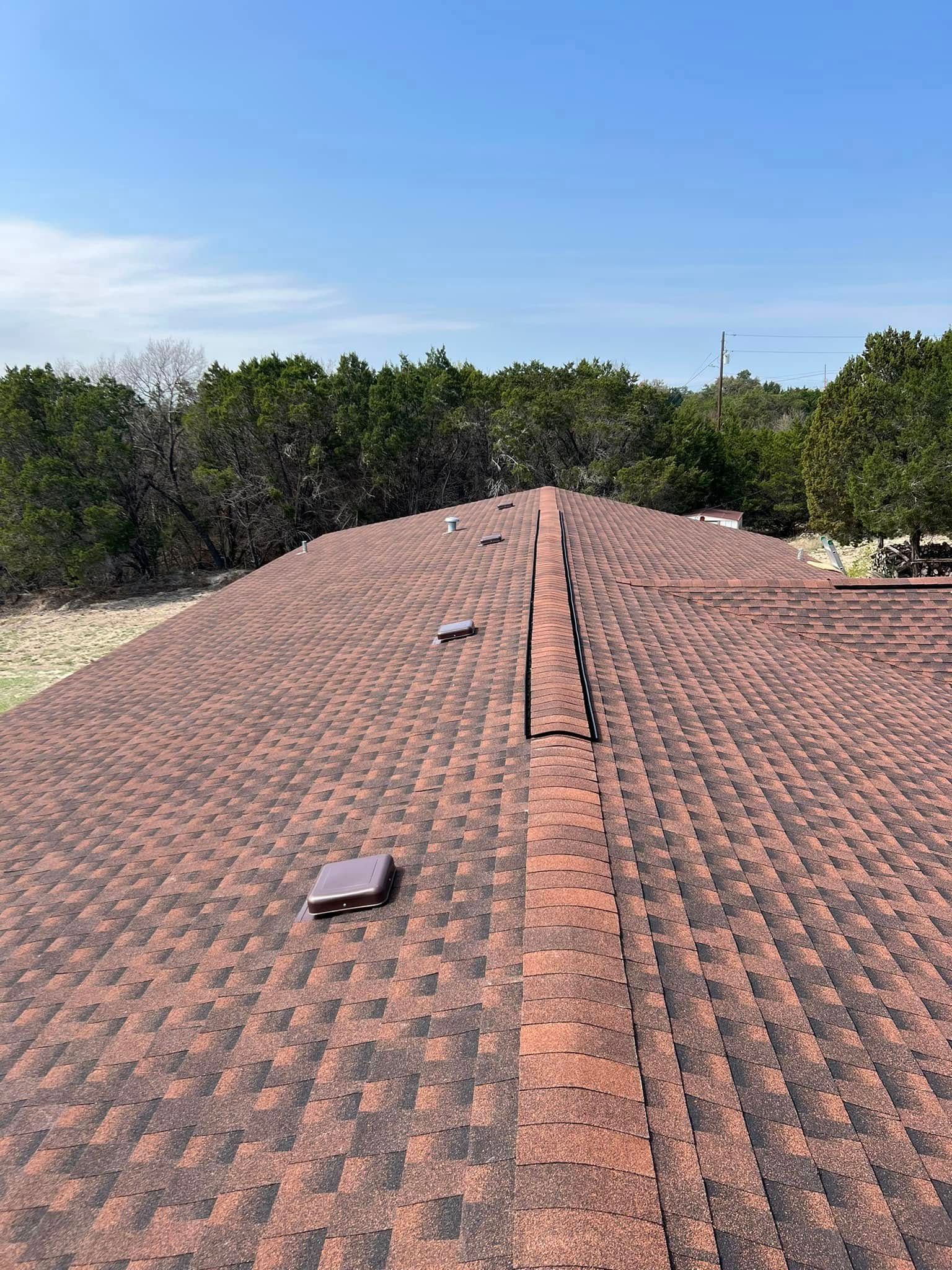 Reddish-brown shingled roof stretching toward trees under a clear blue sky