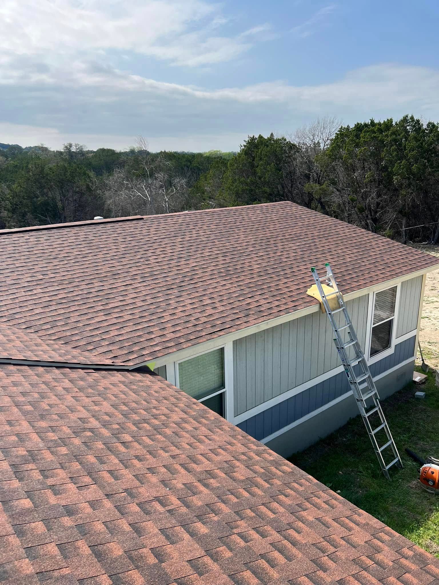 Roofing worker on a ladder inspecting a tan shingle roof beside a house with blue siding
