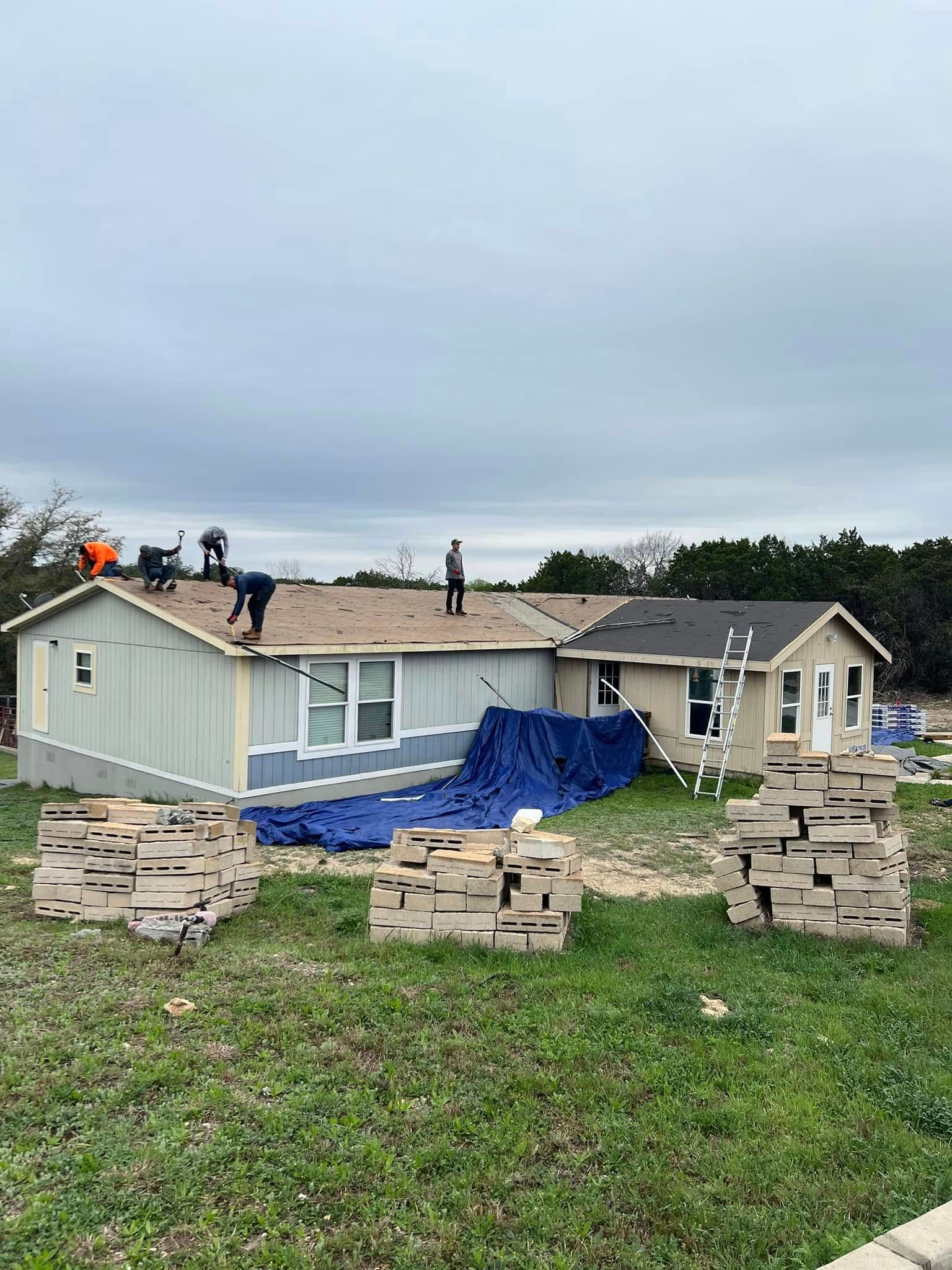Roof being repaired on a house with workers, blue tarp, and stacked bricks in the yard.