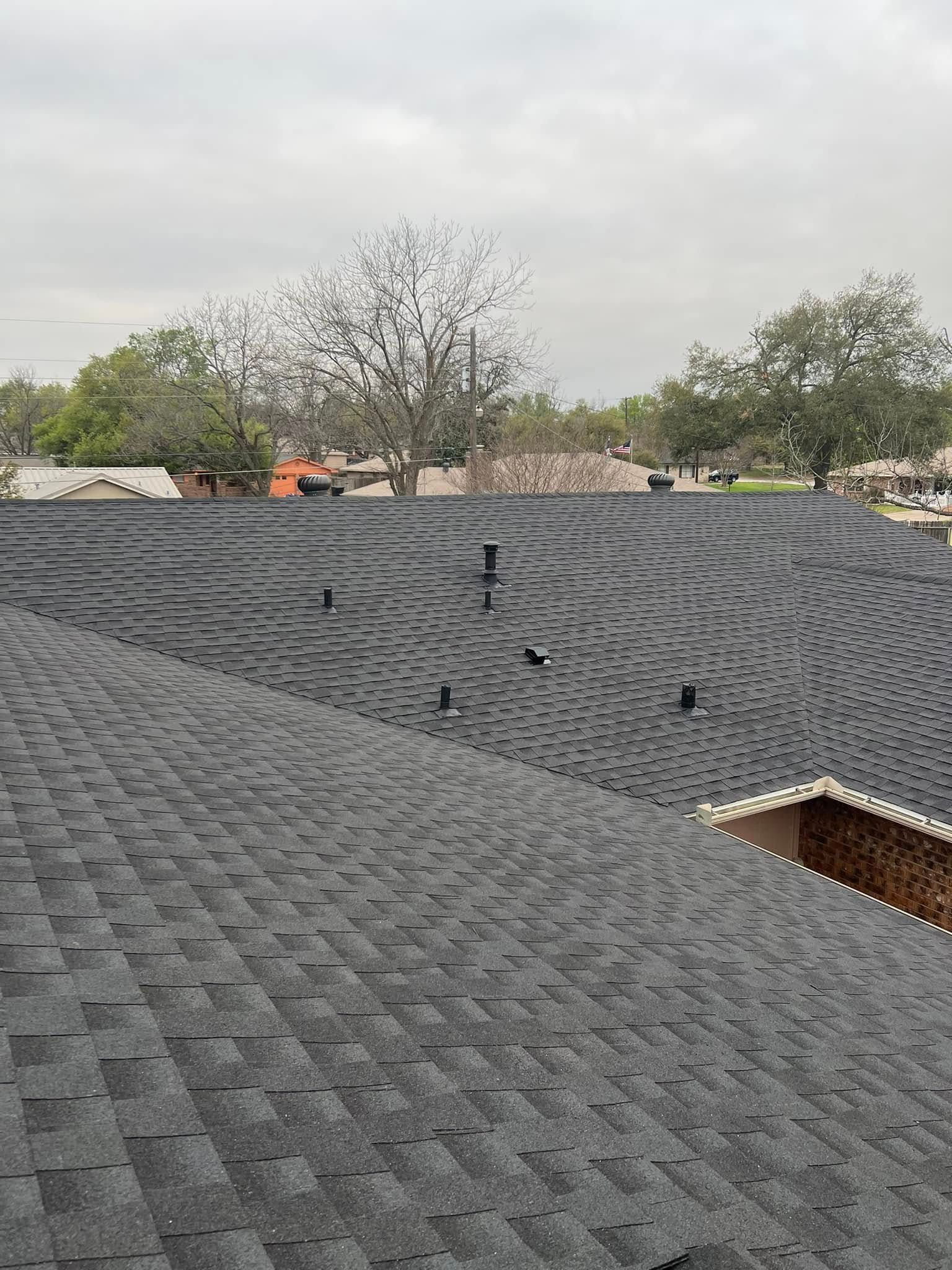 Gray shingled rooftop with vents under an overcast sky, viewed over a neighborhood.
