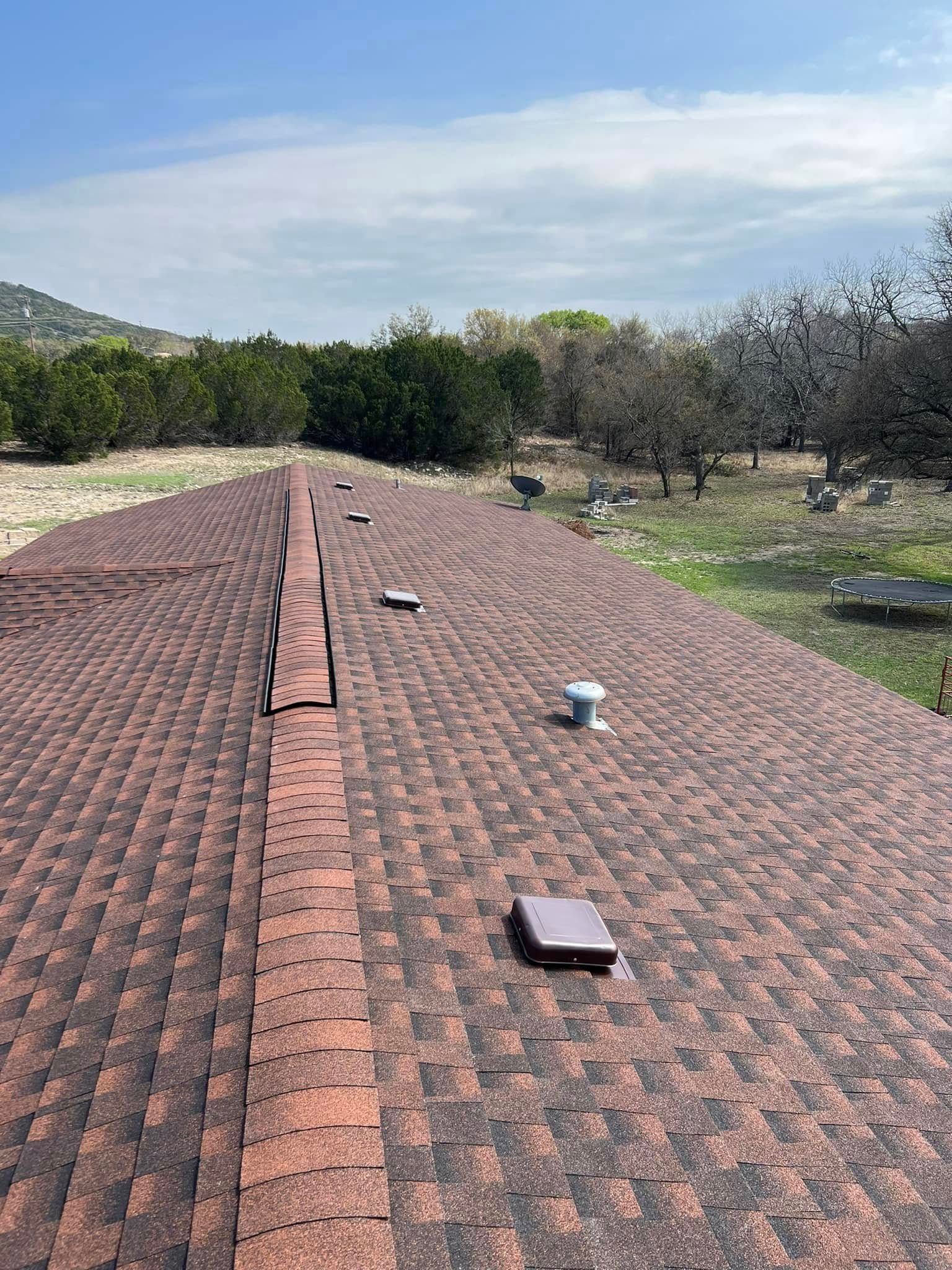 Rooftop with reddish-brown shingles and several vents under a partly cloudy sky.