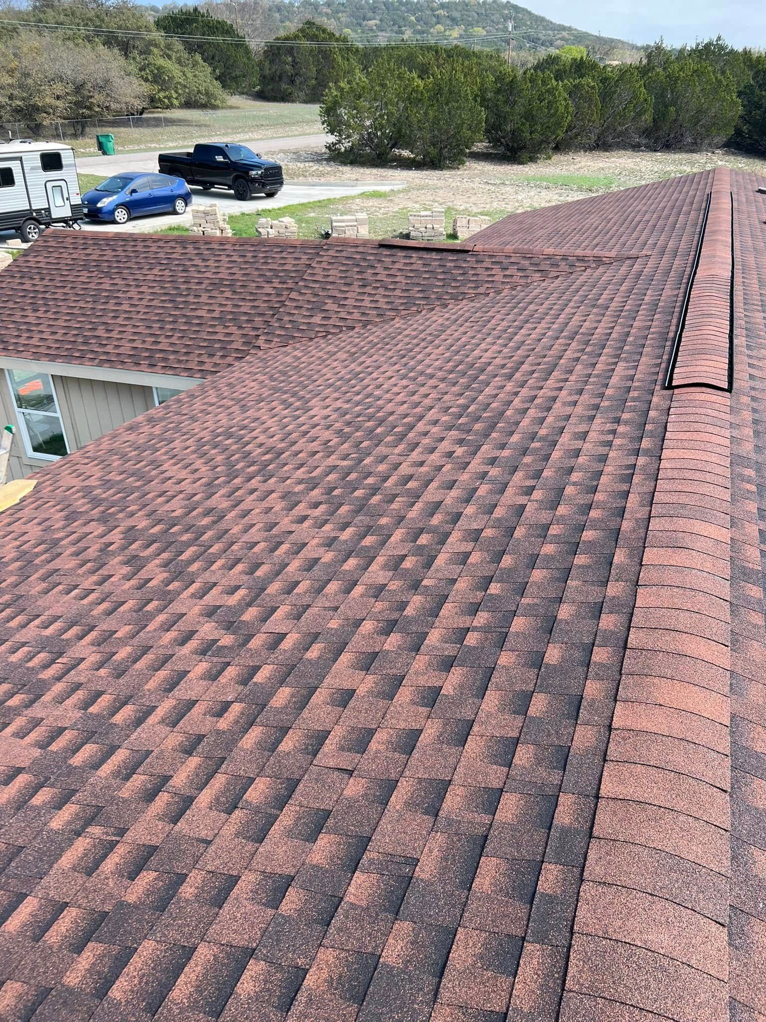 Reddish-brown shingled roof with ridgeline, viewed from above with trees, hills, and parked cars beyond