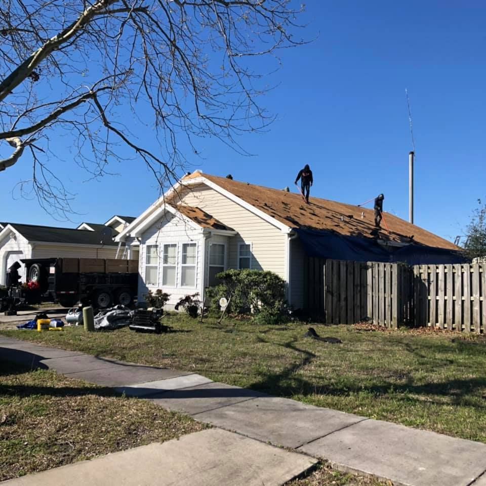 Workers reroofing a beige house under a clear blue sky, with ladders and debris in the yard