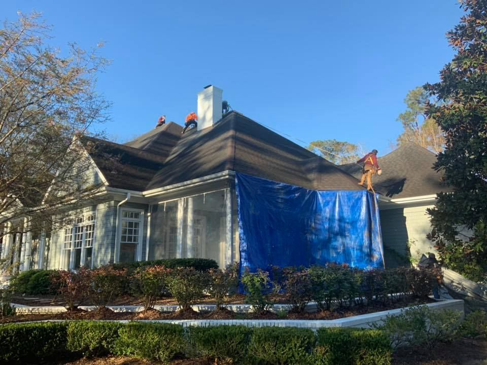 House with blue tarp covering exterior wall under repair, surrounded by trees and shrubs on a sunny day