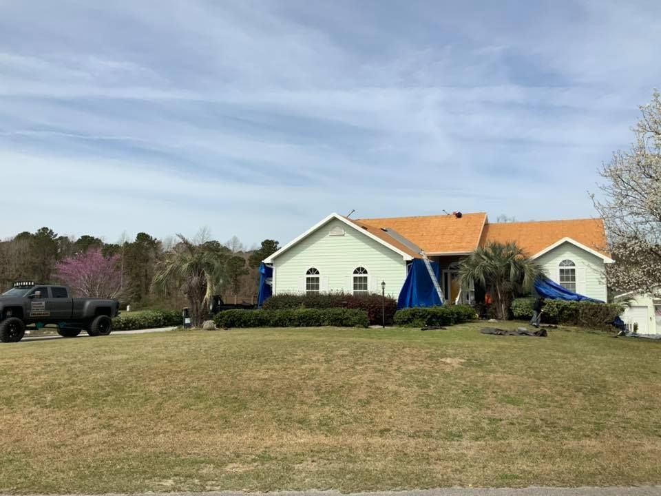Rural house with orange roof and blue awnings behind a grassy field, with a black SUV parked left.