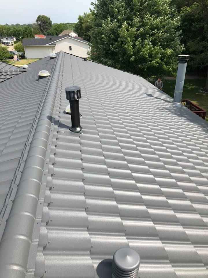Gray metal roof with vents and chimney, viewed from the ridge toward trees and neighboring houses