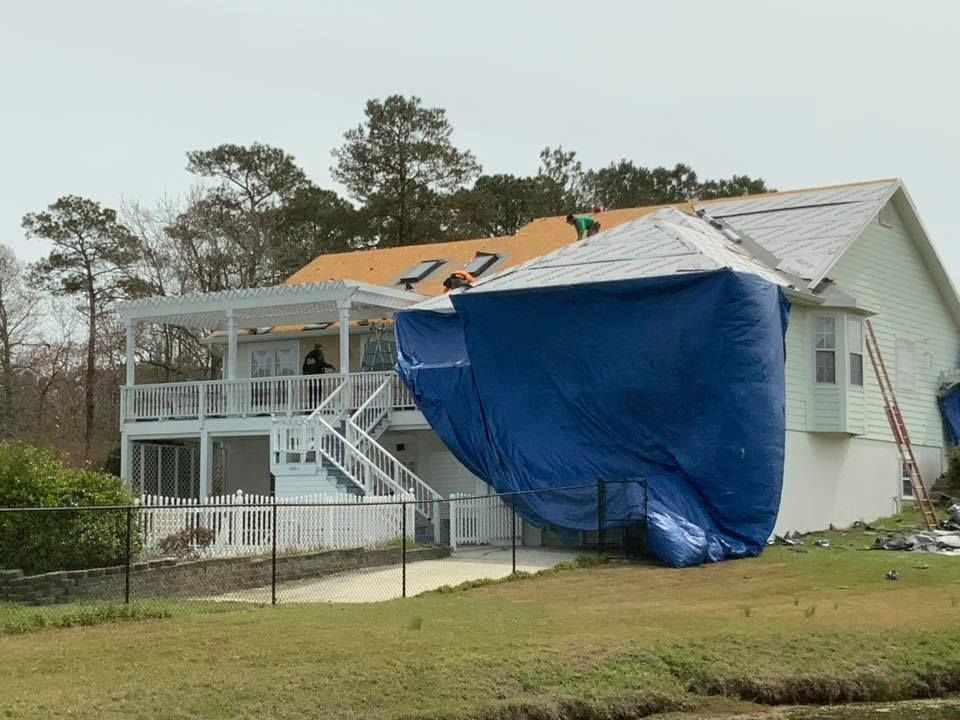 Damaged house with tarp over roof after storm, showing exposed siding and debris in the yard