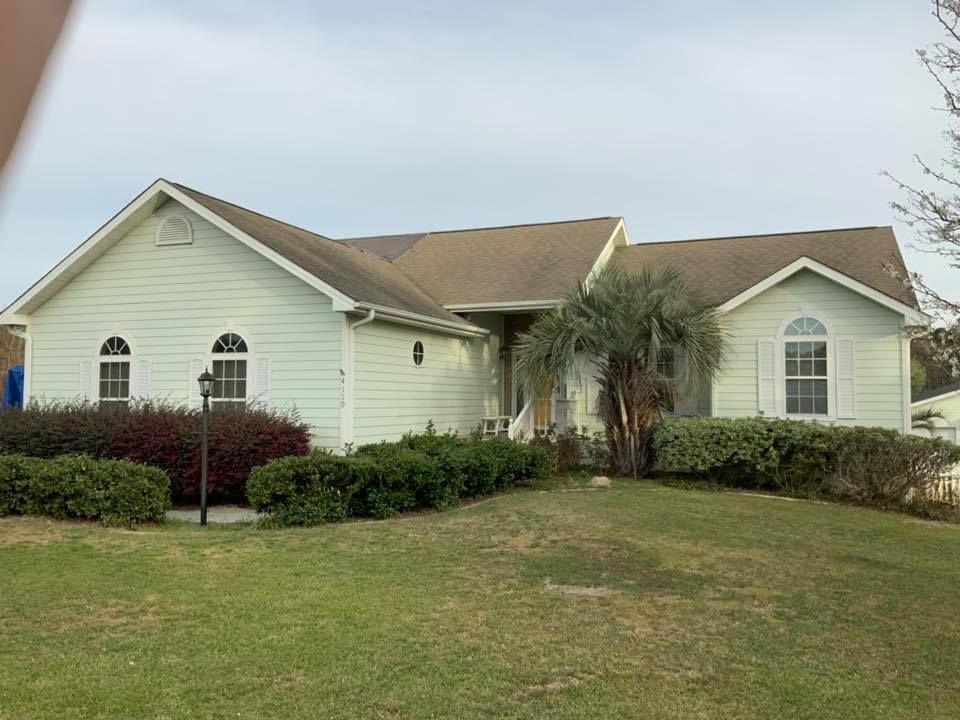 Single-story beige house with white trim, a front lawn, shrubs, and a palm tree under a cloudy sky