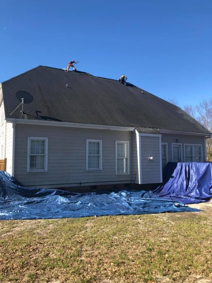 House with a dark roof under repair, with workers on top and blue tarps covering the ground.