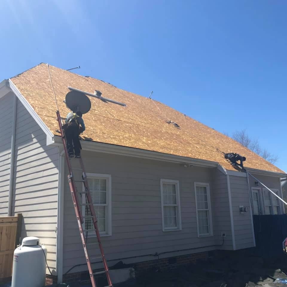 Workers replace a tan shingle roof on a gray house under a clear blue sky.