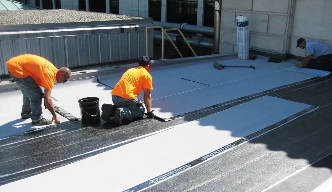 Two workers in orange shirts installing roofing material on a flat roof.