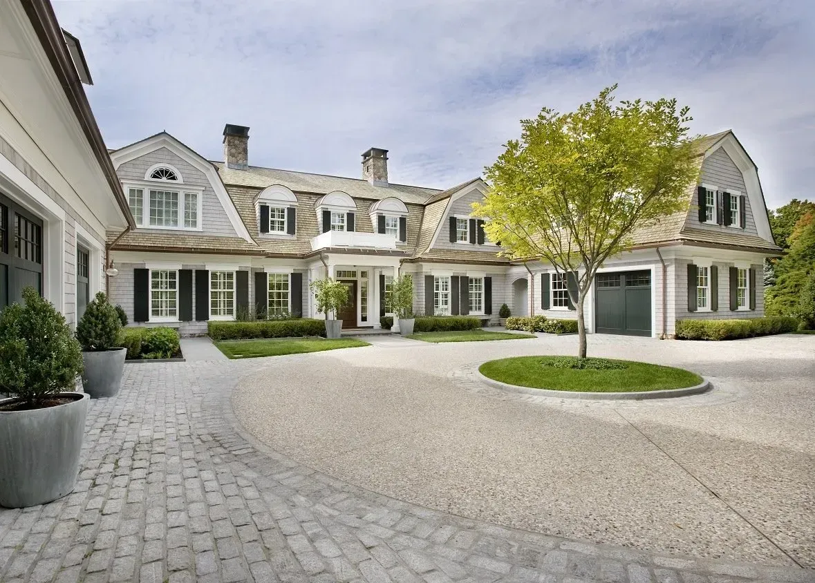 Large gray stone mansion with manicured courtyard, white-trimmed windows, and a central tree under a cloudy sky