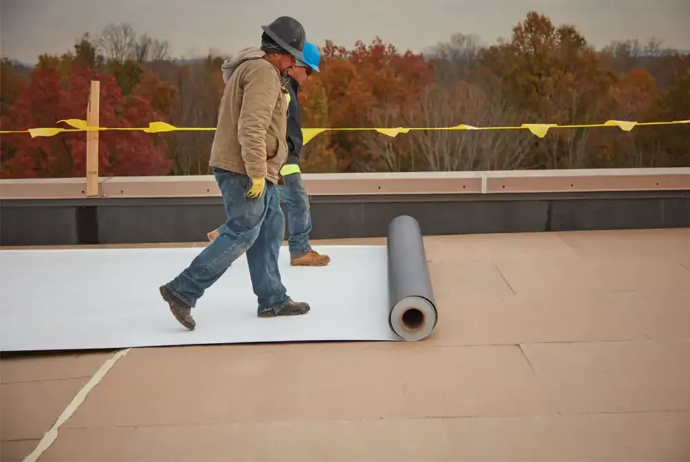 Worker walking on rooftop beside rolled roofing material and yellow safety line
