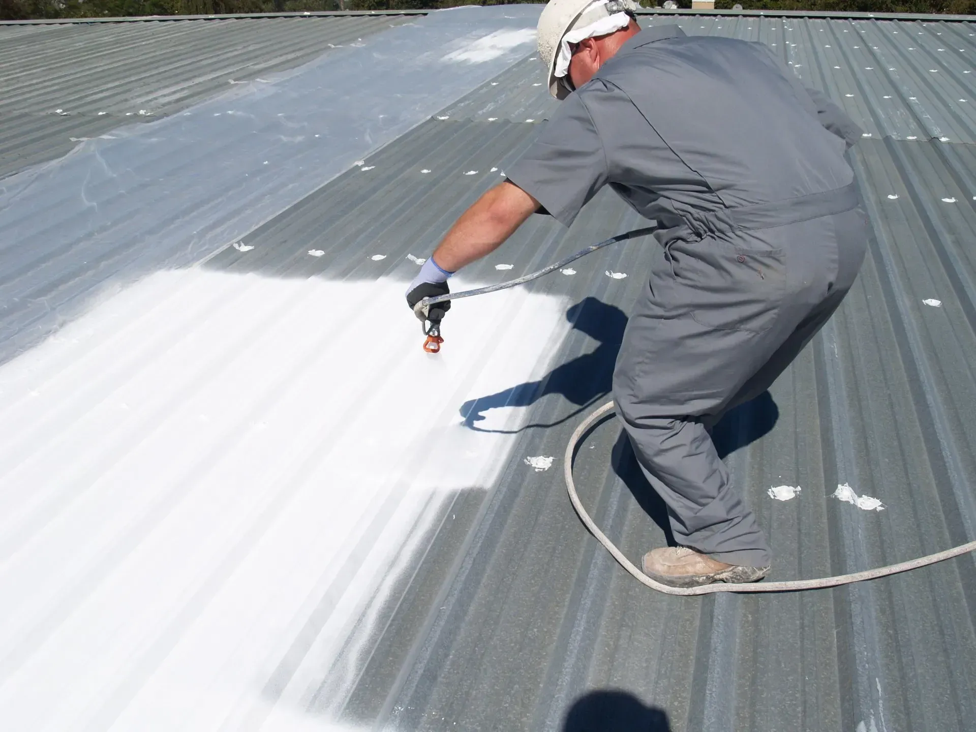 Worker in a hard hat spray-painting a gray metal roof white.