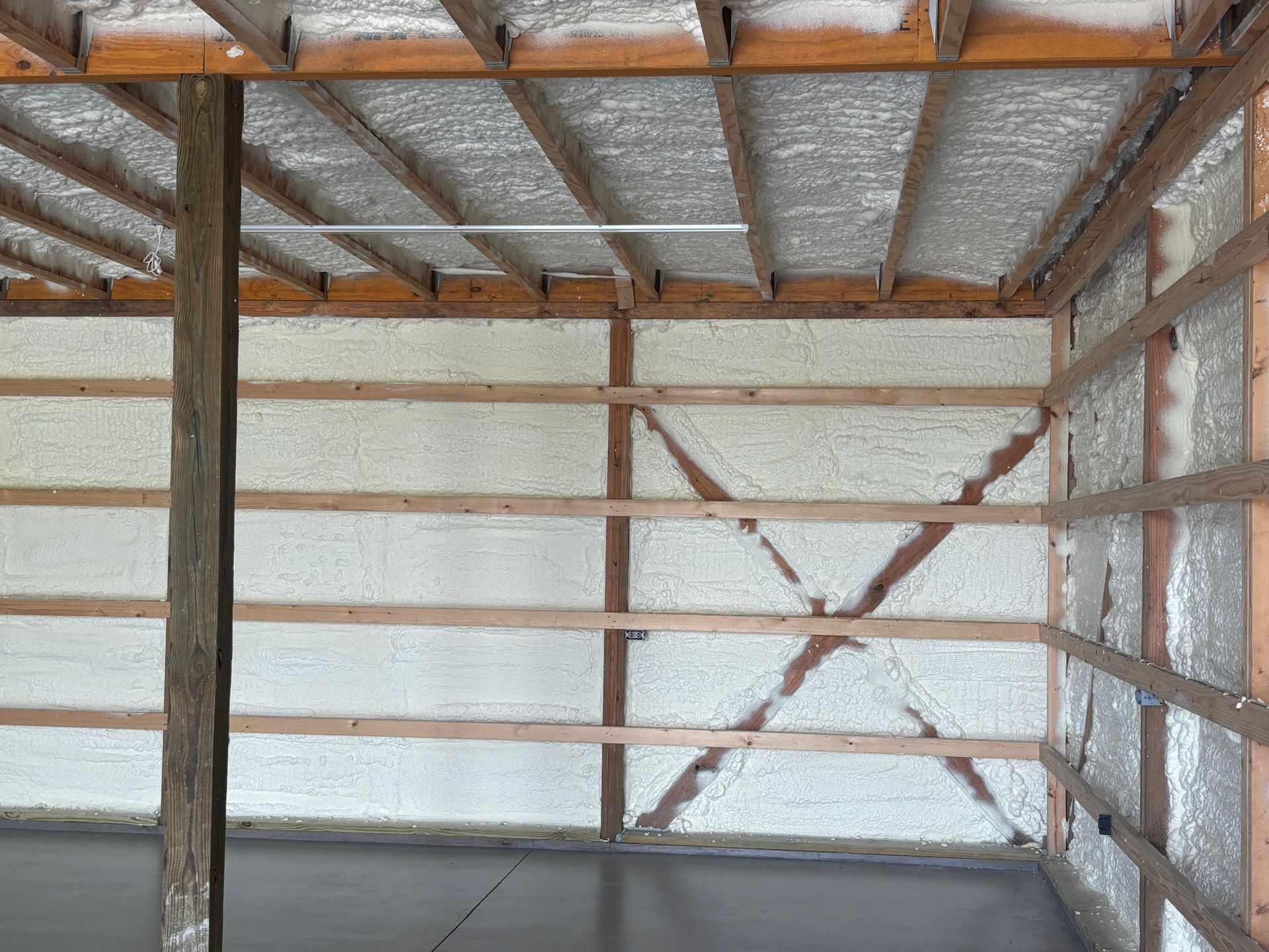 Interior view of a building frame insulated with white spray foam. Wooden beams and a gray concrete floor are visible.