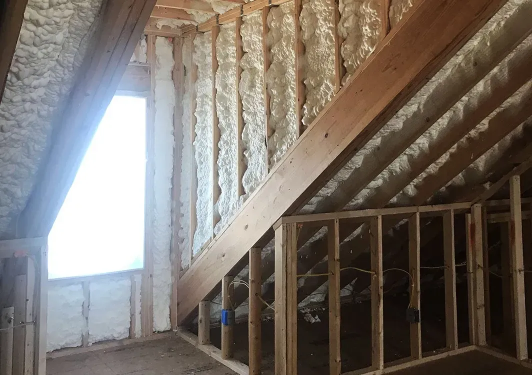 Interior of an attic with spray foam insulation between wooden beams and studs, with a window visible.