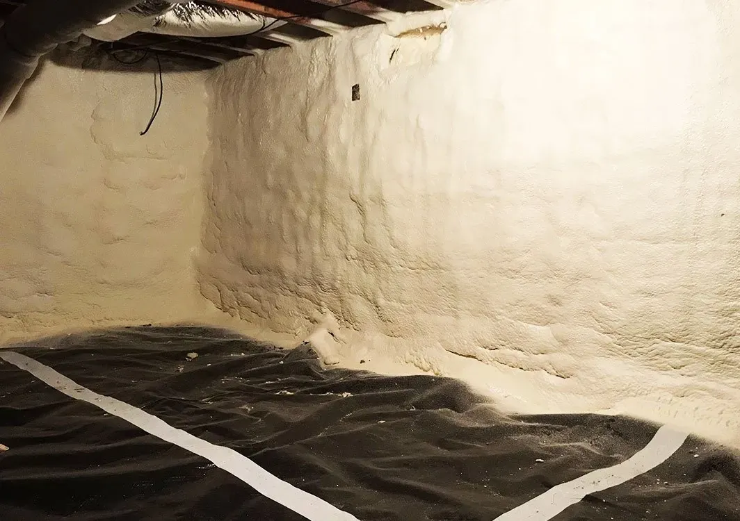 Basement corner with white spray foam insulation on walls, black ground cover, and exposed ceiling.