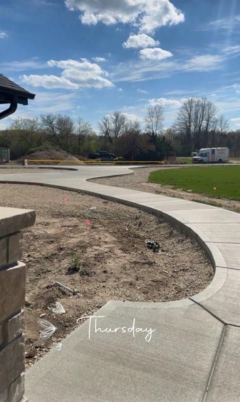 Curved concrete walkway beside a dirt bed and grassy park area under a cloudy blue sky