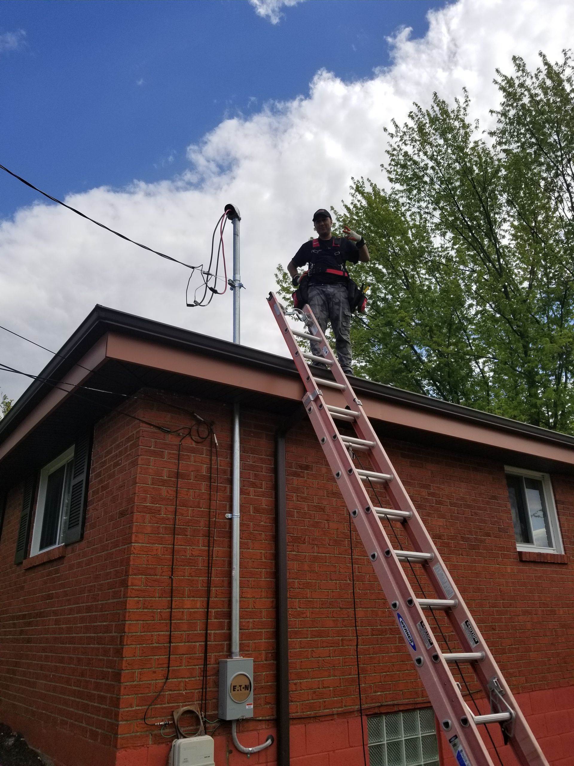 Worker on a ladder inspecting a brick building roof beside a utility pole under a blue sky