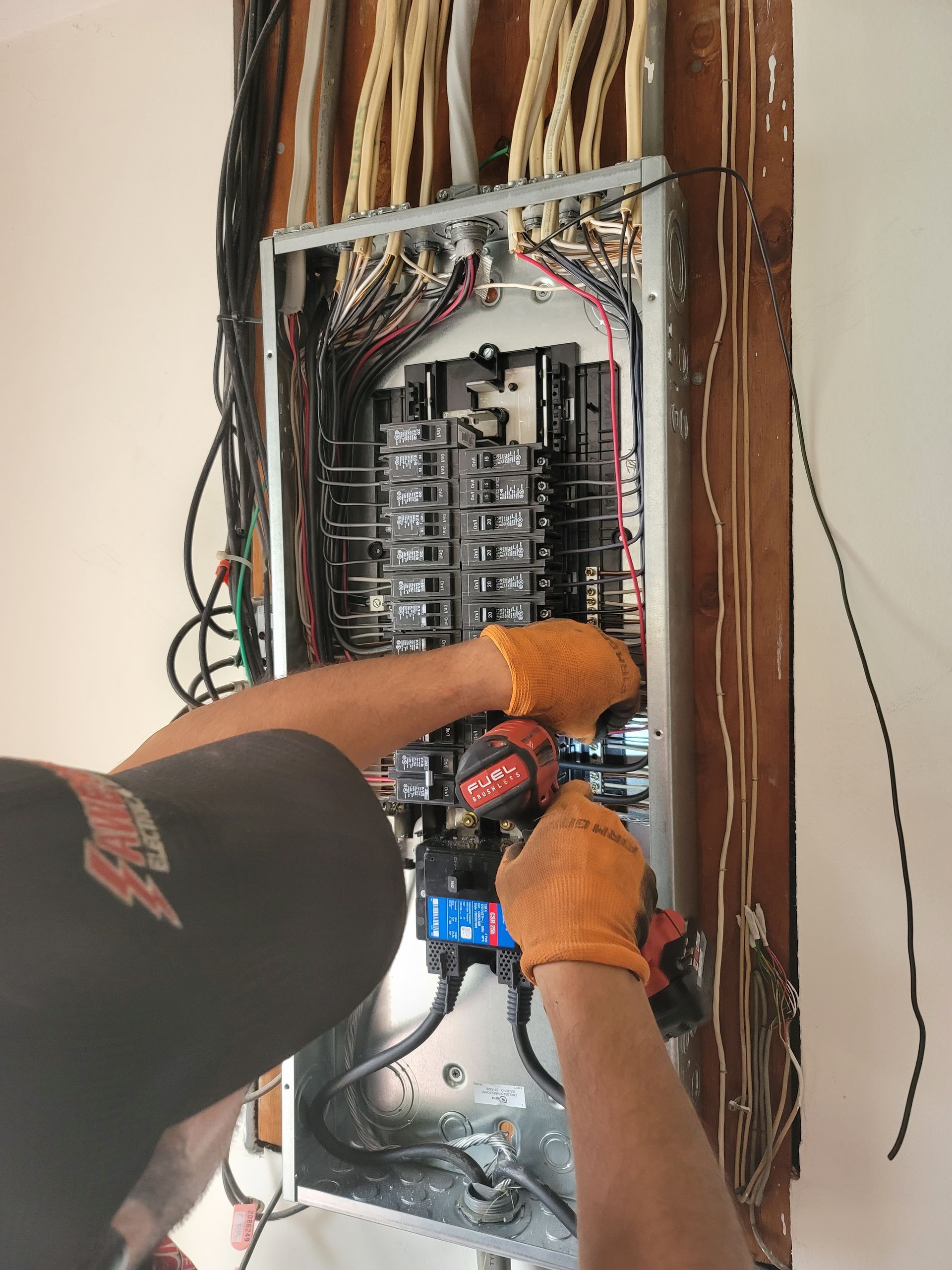 Electrician working inside an open breaker panel with exposed wiring and orange gloves.