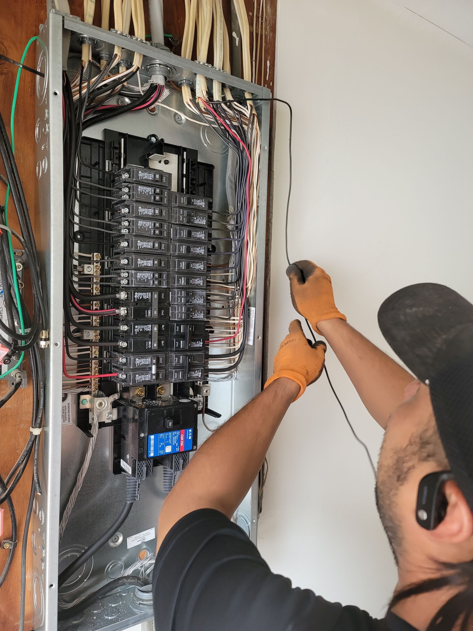 Electrician wiring a breaker panel in an open wall, using a drill beside exposed electrical panels.