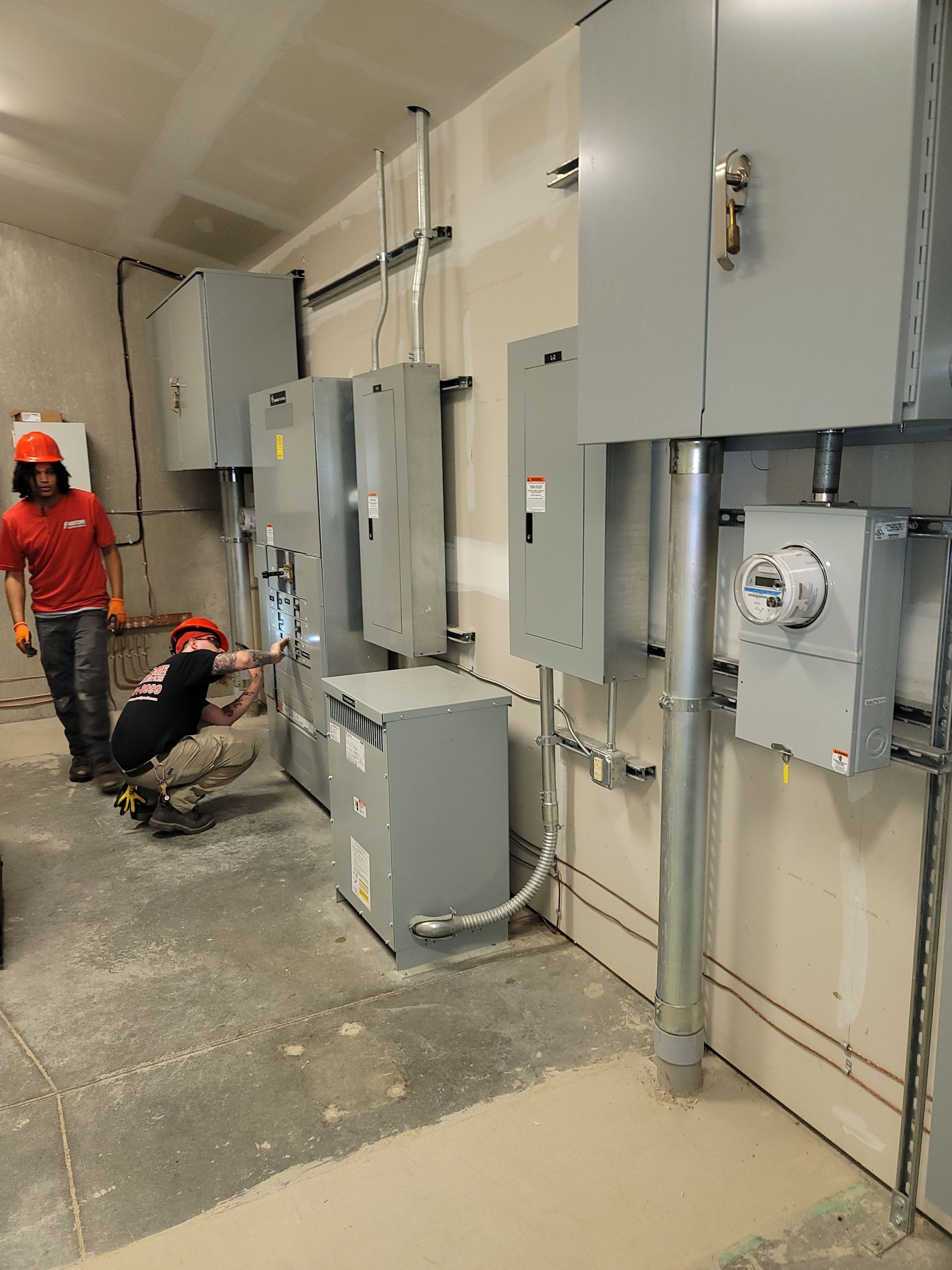 Workers in red uniforms inspect electrical panels in an industrial hallway.