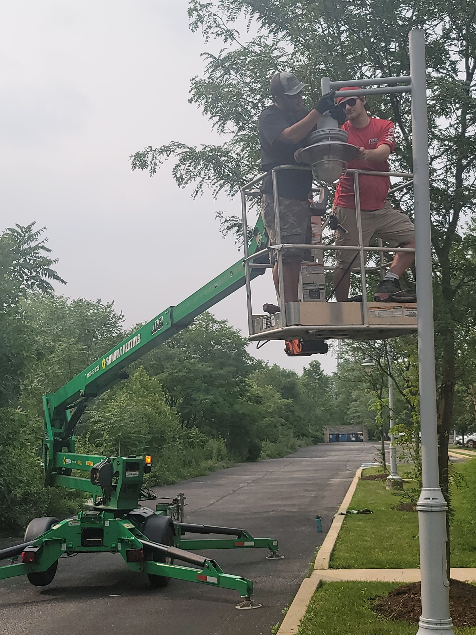 Workers in a lift trimming a tree beside a roadside, with a green cherry picker below.