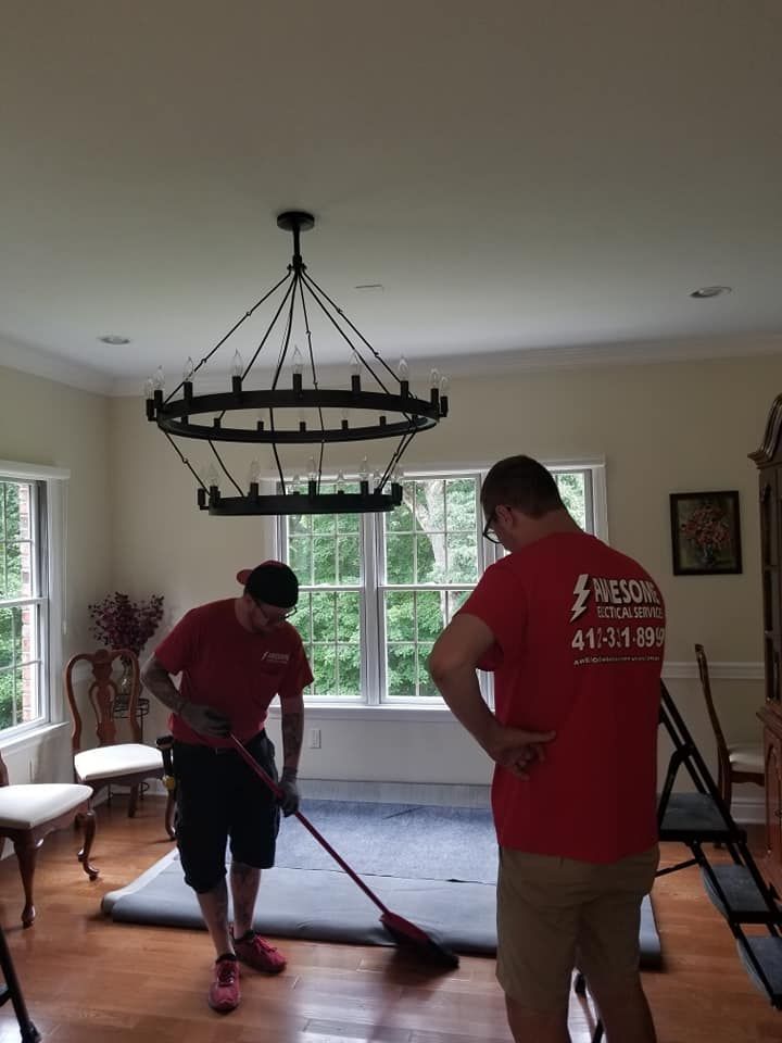 Two people mopping a hardwood floor in a sunlit living room with a large chandelier.