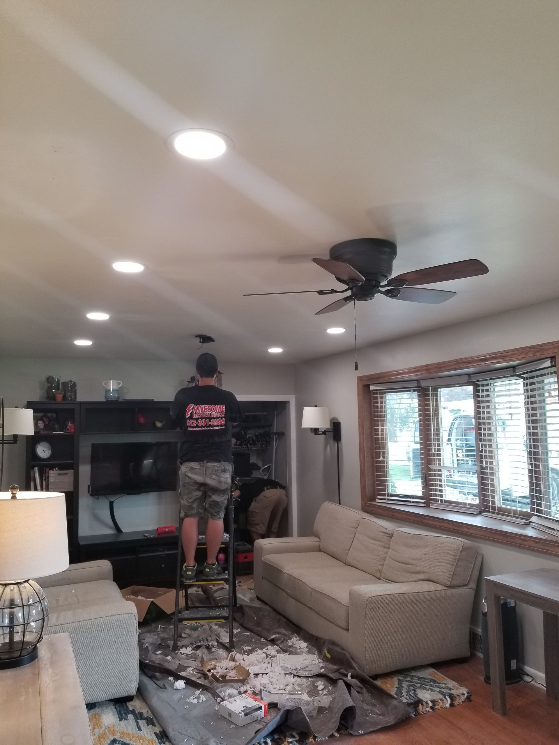 Person on a step ladder fixing a ceiling fan in a living room with a beige couch and windows.
