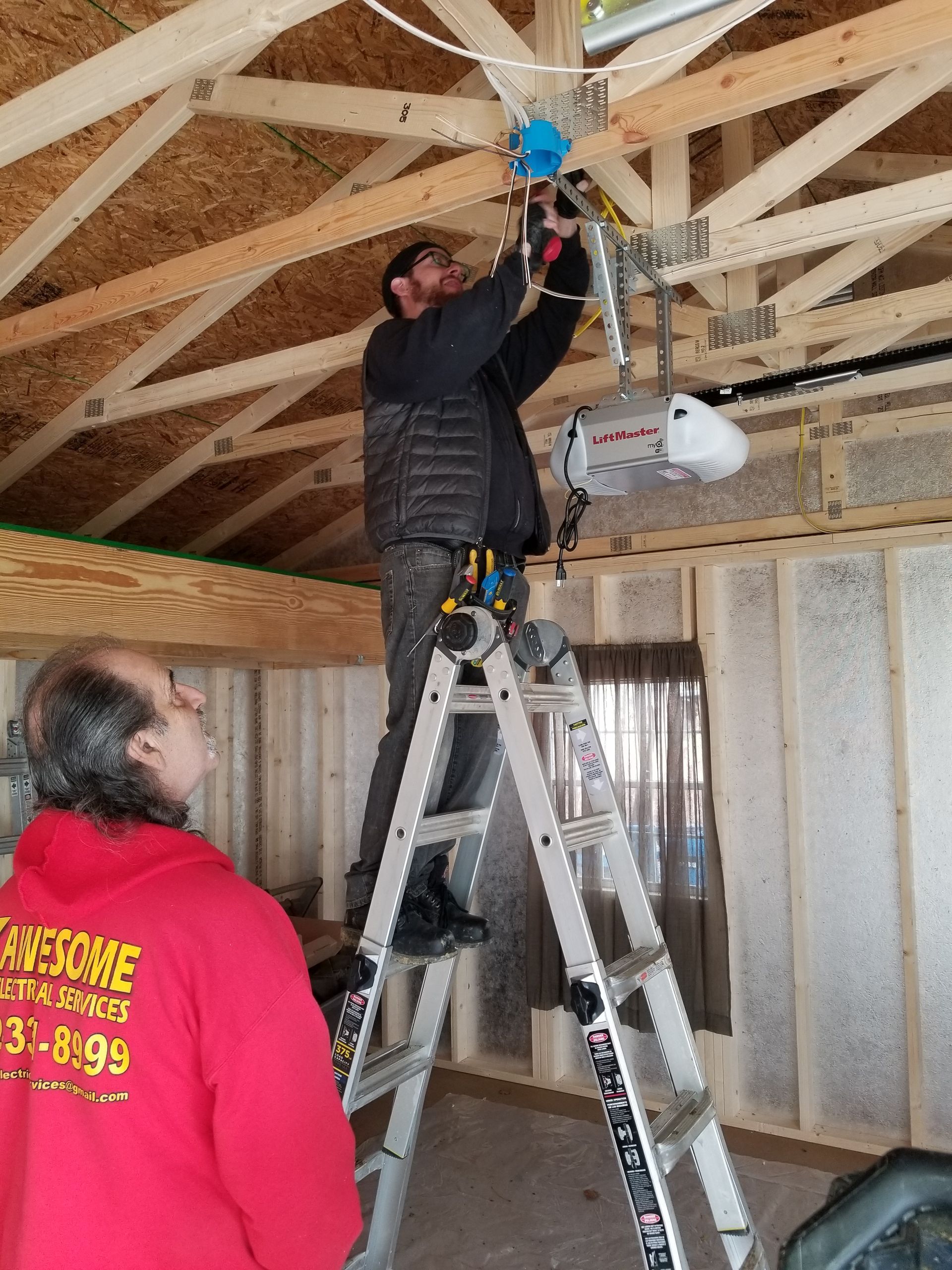 Two workers wiring a ceiling fan in an unfinished room, one on a ladder and one watching nearby.