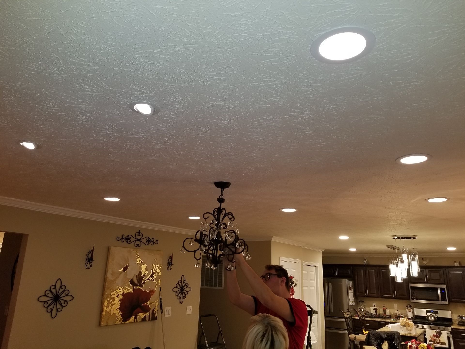 Person hanging a chandelier in a beige kitchen with recessed ceiling lights and wall decor